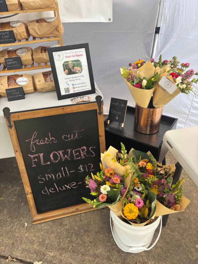 A display at a farmer's market showing bouquets of locally grown, fresh-cut flowers from Brighter Day Flower Farm for sale next to Sota Dough bread. A chalkboard sign lists flower prices, and a framed informational sign features a photo of the flower farmer.